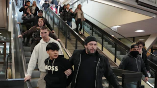A plainclothes, unmasked federal agent holds on to a man as they go down the escalator. Other federal agents stand nearby, and onlookers record the incident on their phone.