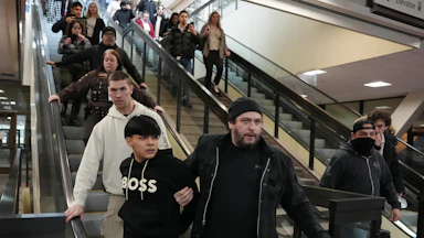 A plainclothes, unmasked federal agent holds on to a man as they go down the escalator. Other federal agents stand nearby, and onlookers record the incident on their phone.