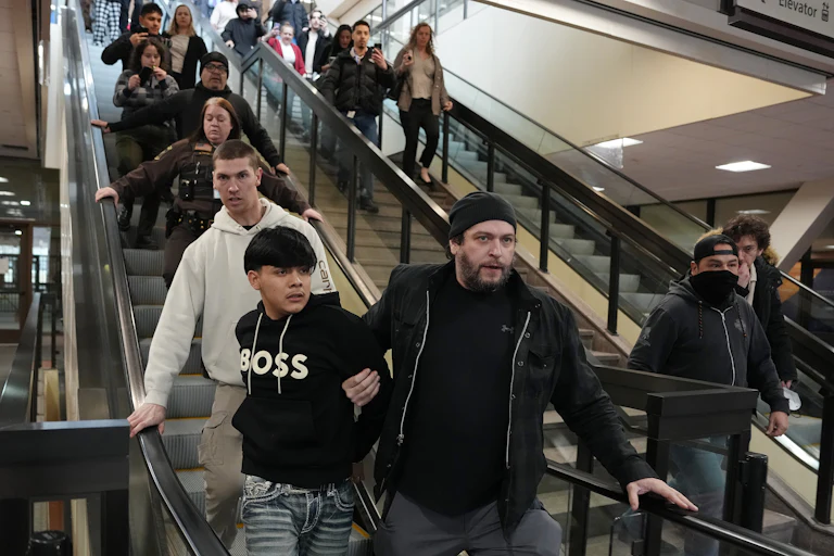 A plainclothes, unmasked federal agent holds on to a man as they go down the escalator. Other federal agents stand nearby, and onlookers record the incident on their phone.