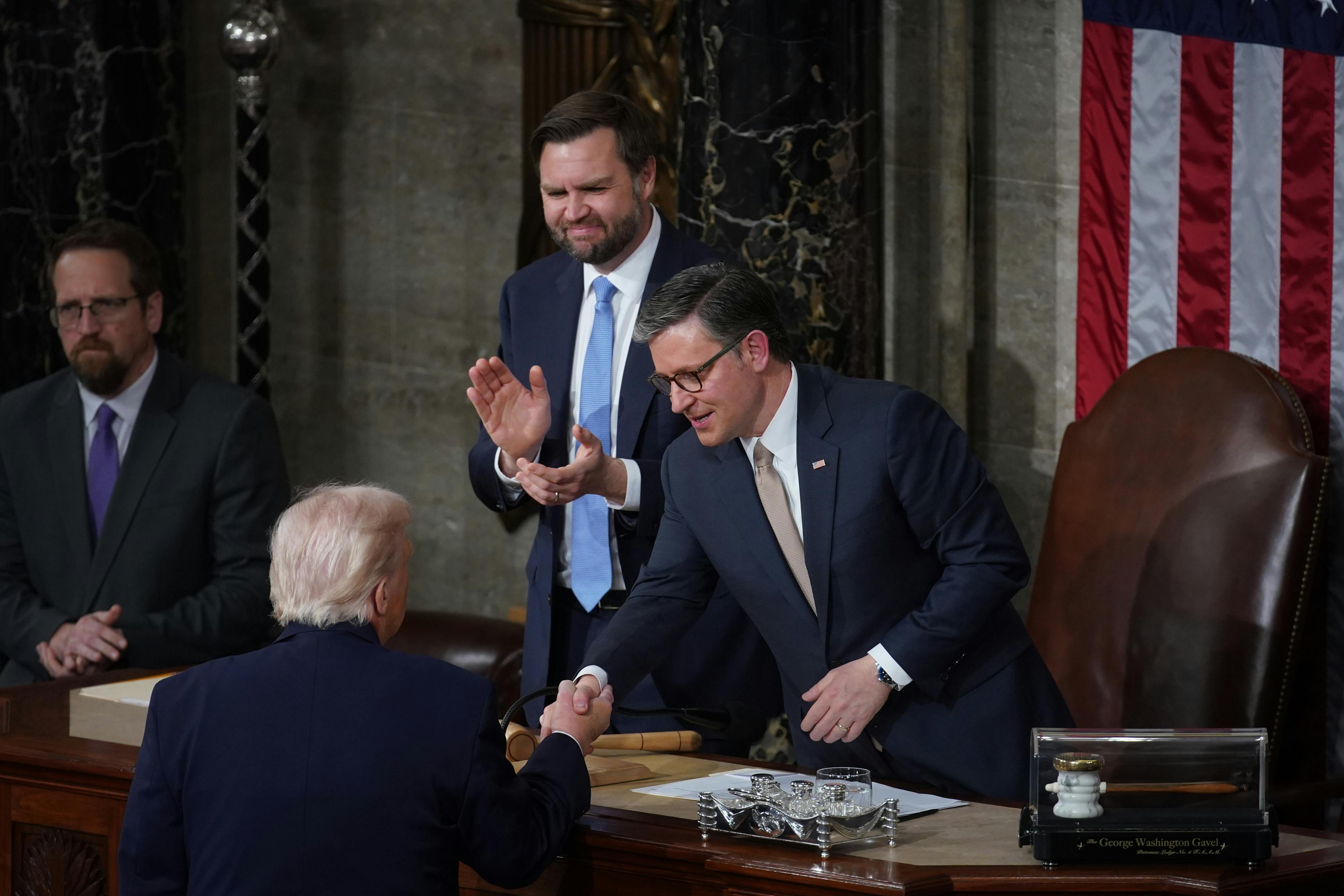 House Speaker Mike Johnson shakes hands with Donald Trump at the lectern after the State of the Union. Vice President JD Vance stands next to Johnson and claps