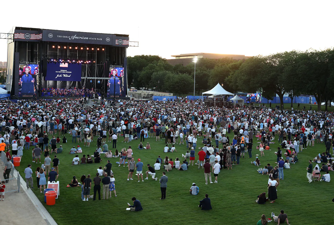 People stand and sit on the grass during a speech at the Great American State Fair