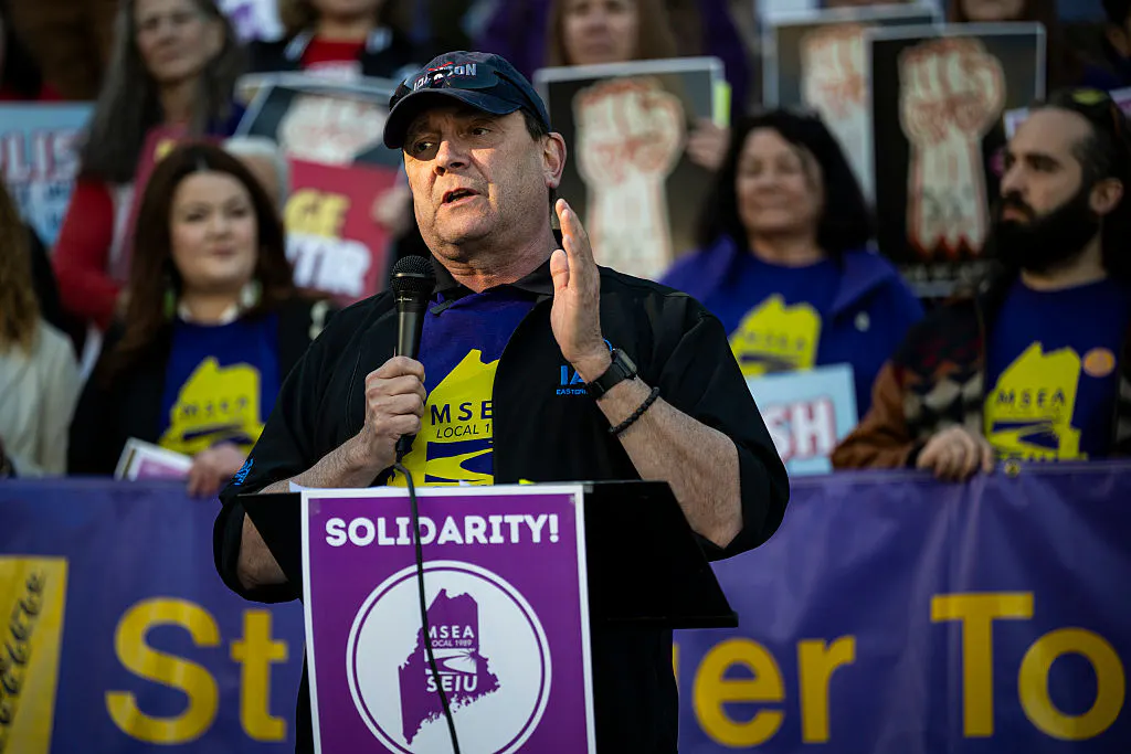 Troy Jackson holds a microphone in front of a lectern saying "Solidarity!" Numerous supporters stand behind him.