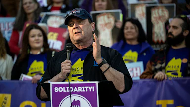 Troy Jackson holds a microphone in front of a lectern saying "Solidarity!" Numerous supporters stand behind him.