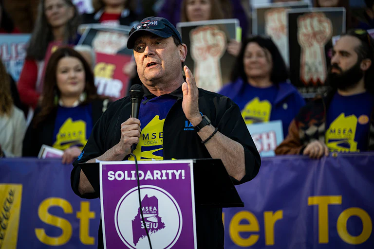 Troy Jackson holds a microphone in front of a lectern saying "Solidarity!" Numerous supporters stand behind him.