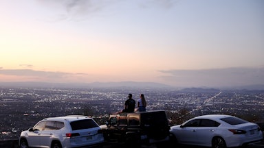 Two people sit on the top of a car at sunset looking out over a city. 