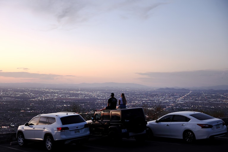 Two people sit on the top of a car at sunset looking out over a city. 