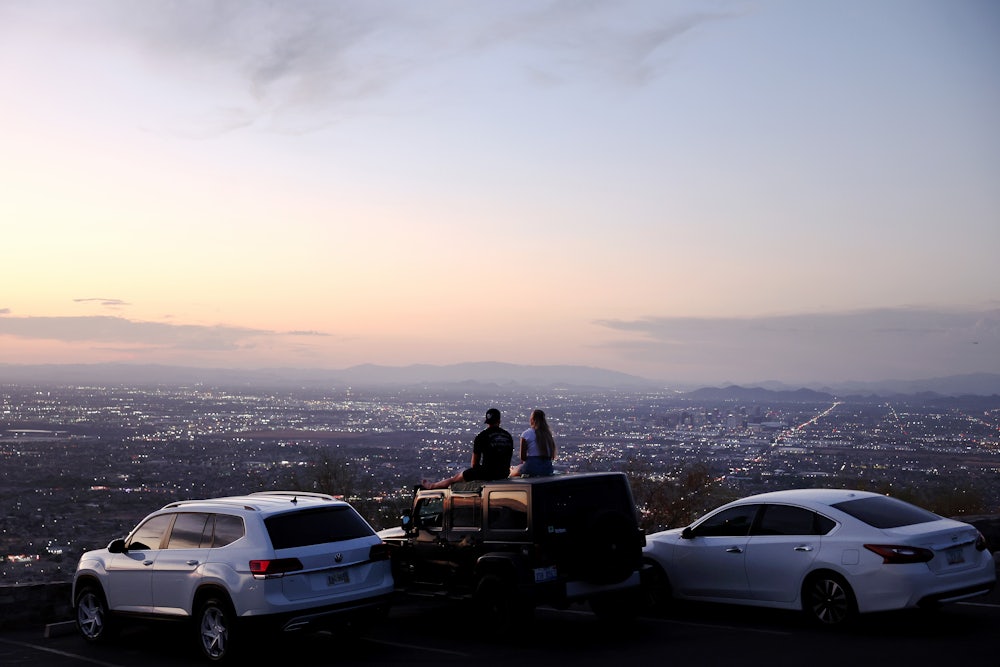Two people sit on the top of a car at sunset looking out over a city.