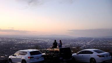 Two people sit on the top of a car at sunset looking out over a city.