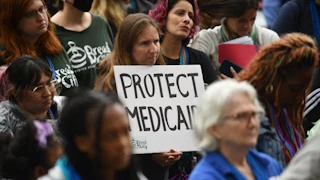 A crowd of protesters stands while one woman in the middle holds a handwritten "PROTECT MEDICAID" sign.