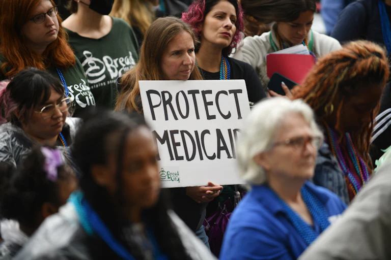 A crowd of protesters stands while one woman in the middle holds a handwritten "PROTECT MEDICAID" sign.