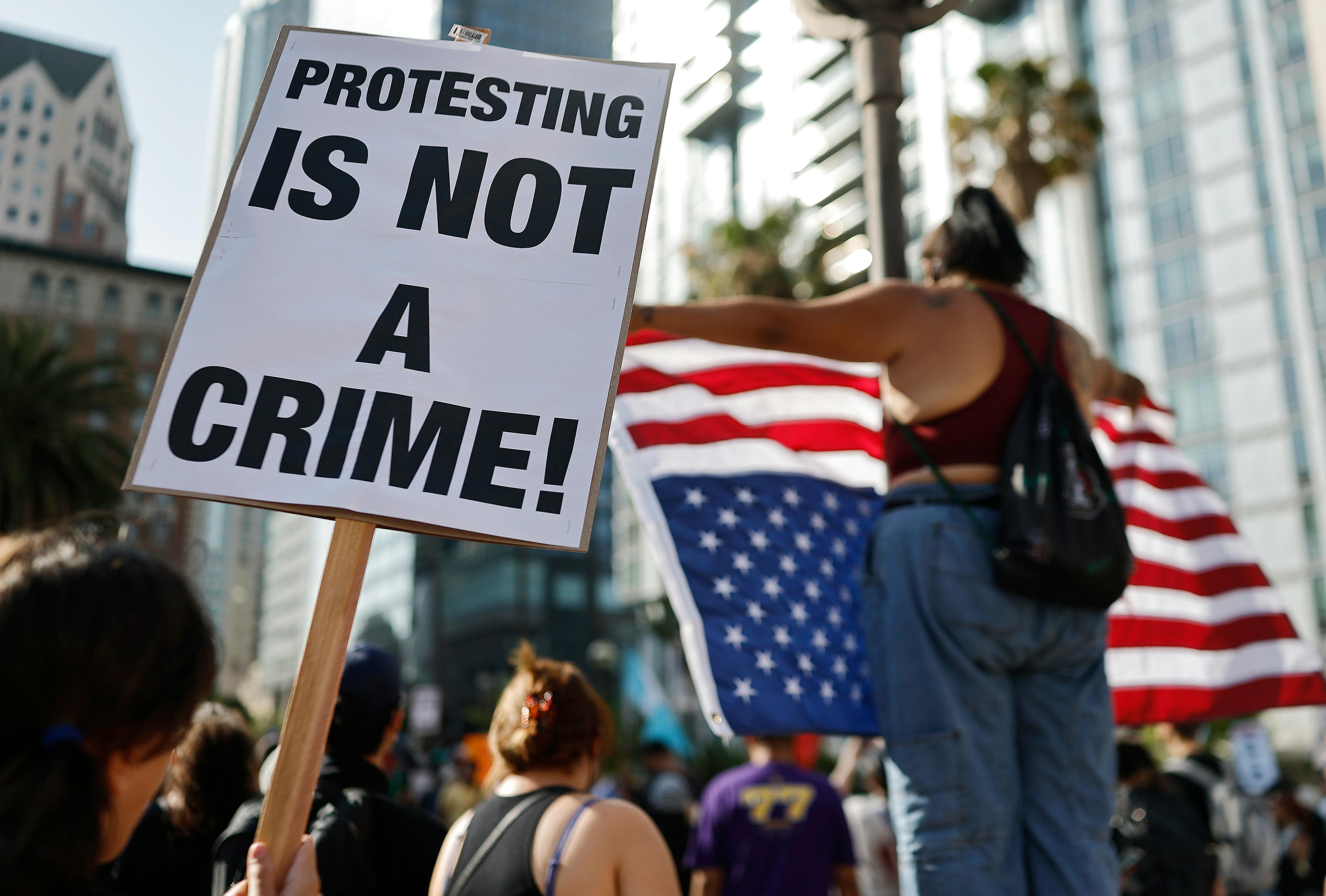 someone holds a sign reading protesting is not a crime sign while another holds up an upside down American flag 