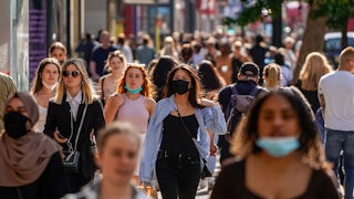 A crowd of people, some masked and some unmasked, walk down a busy street.