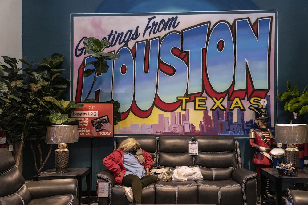 A masked woman sits on a couch at a furniture store turned warming station in front of a large sign reading “Greetings from Houston Texas”