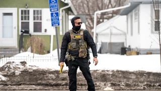 An ICE agent holds a taser while standing on the street, wearing a face mask.