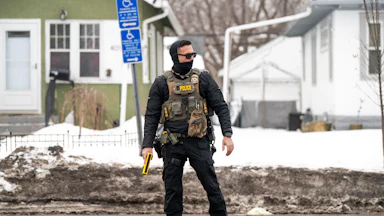 An ICE agent holds a taser while standing on the street, wearing a face mask.