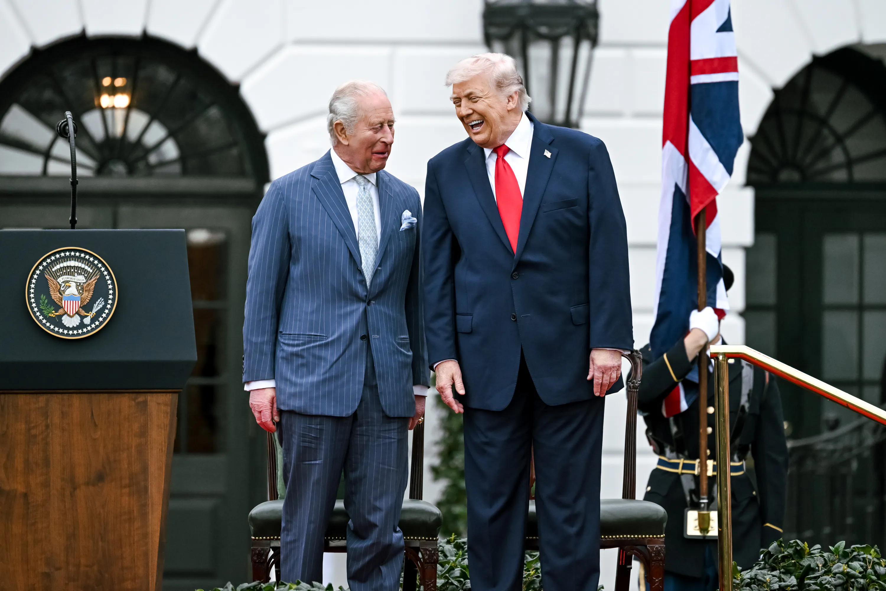 King Charles III and Donald Trump laugh while standing next to each other on a platform outside the White House