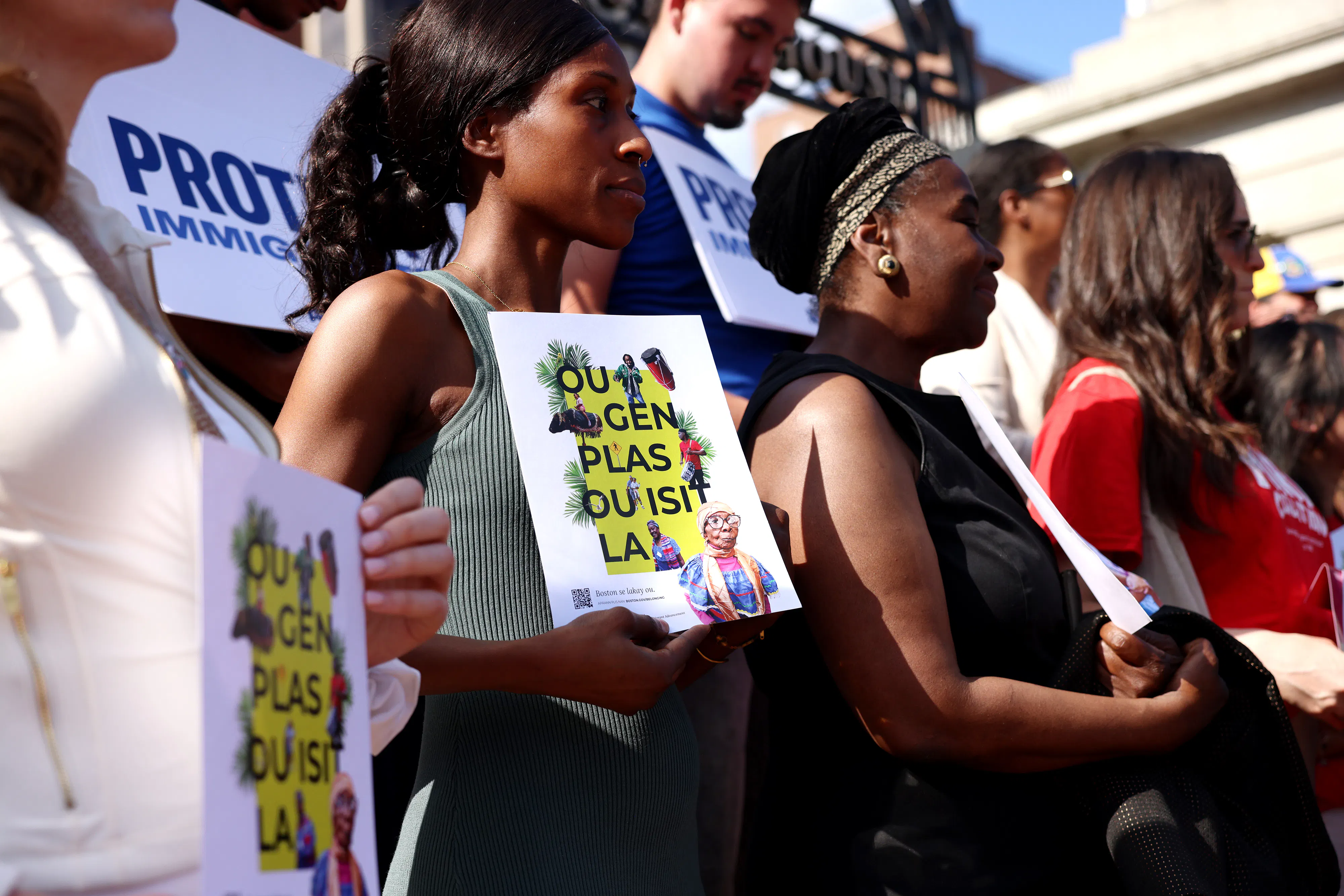 Community members hold signs of support written in Haitian Creole that translate to “You Belong Here” during a press conference held in the wake of the Supreme Court decision to overturn TPS for Haitians and Syrians outside of the State House on June 25, 2026. 
