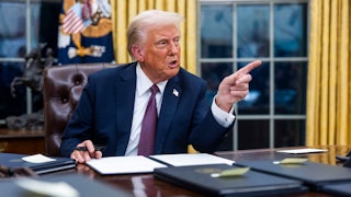 Donald Trump points while speaking, as he sits at his desk with executives orders in front of him in black folders.