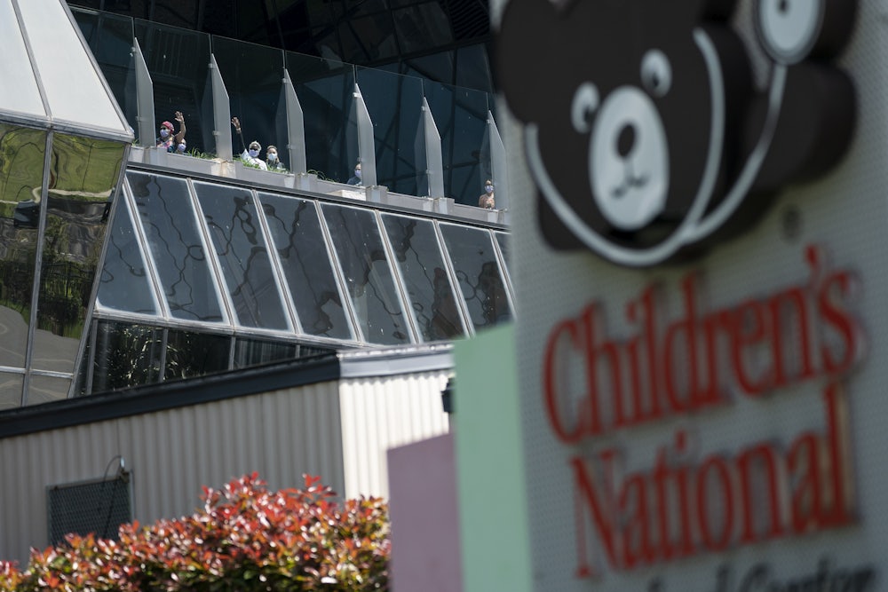 This photo shows the exterior of Children's National Hospital, including a sign with their bear logo. People stand at the windows.