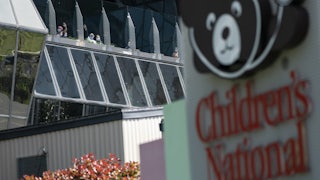 This photo shows the exterior of Children's National Hospital, including a sign with their bear logo. People stand at the windows.