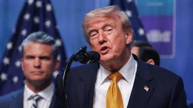 Donald Trump speaks at a podium during a NATO summit. Defense Secretary Pete Hegseth stands behind him.