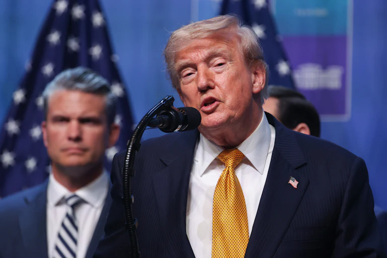 Donald Trump speaks at a podium during a NATO summit. Defense Secretary Pete Hegseth stands behind him.