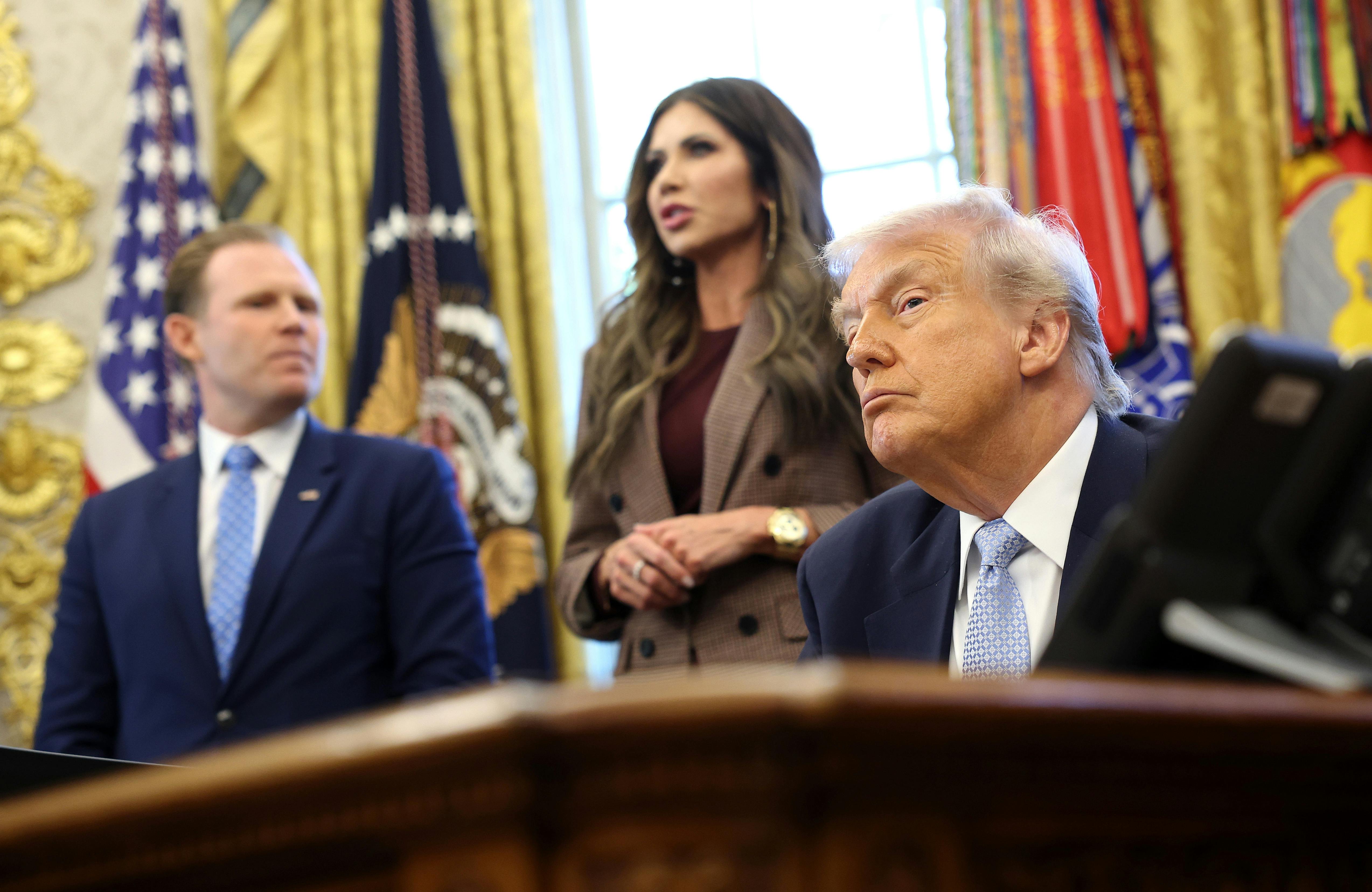 Donald Trump sits at the desk in the Oval Office, with Kristi Noem and Andrew Giuliani behind him.