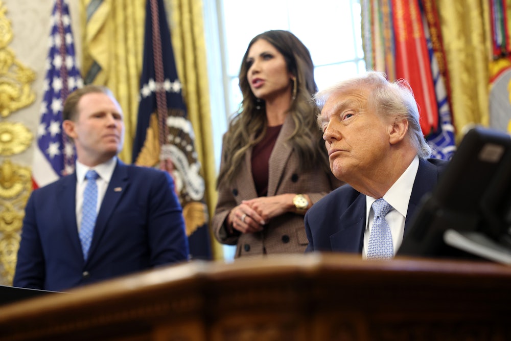 Donald Trump sits at the desk in the Oval Office, with Kristi Noem and Andrew Giuliani behind him.