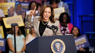 Harris points one finger skyward while smiling at a podium with fans in the background.