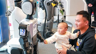 An adult holds a baby who reaches out to hold hands with a humanoid robot.