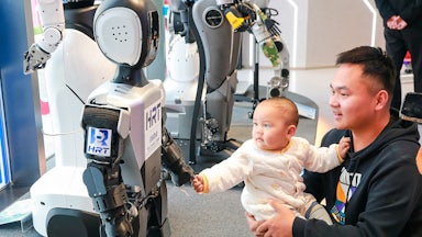 An adult holds a baby who reaches out to hold hands with a humanoid robot.