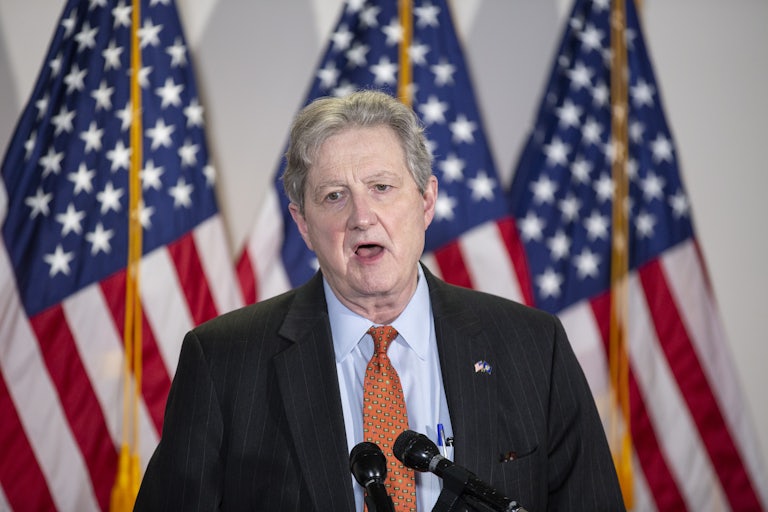 Senator John Kennedy opens his mouth and stares off into the distance while standing at a lectern. Three U.S. flags are behind him.