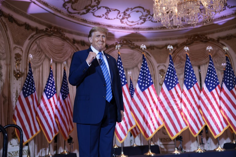 Donald Trump smiles weirdly and makes a fist. A row of U.S. flags are behind him, as are massive drapes, and a huge chandelier can be seen above him. He is at Mar-a-Lago.