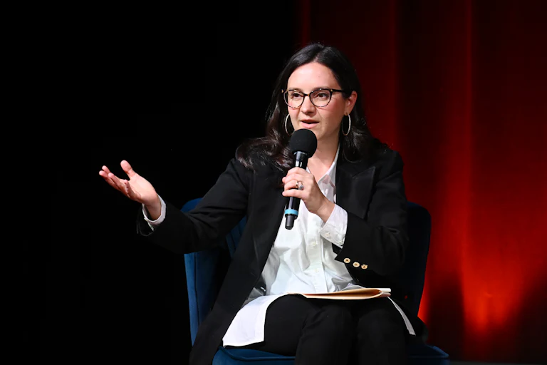 Bari Weiss gestures and speaks while sitting during an event. She holds a microphone in one hand.