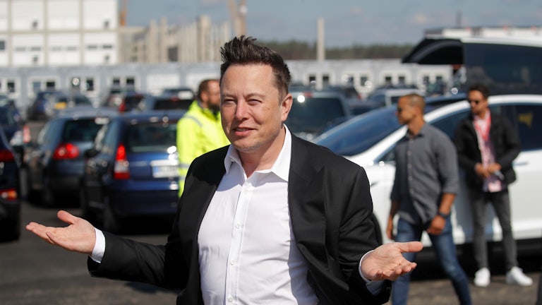 Elon Musk, standing before a fleet of cars, with hair and jacket swept up by the breeze 