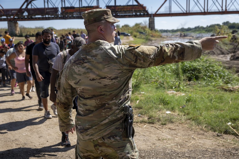 A person in camouflage uniform points off camera with a line of people in front of him.