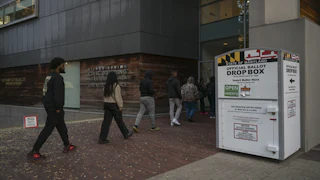 A line of people walk into a polling place labeled "civic building" past an official ballot drop box with the Maryland flag on it.