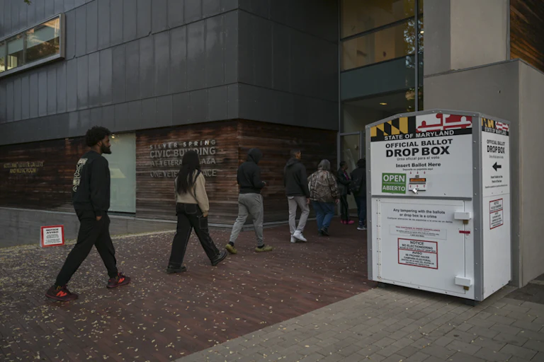 A line of people walk into a polling place labeled "civic building" past an official ballot drop box with the Maryland flag on it.