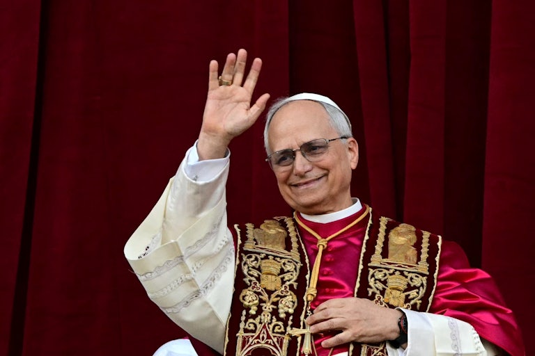 Pope Leo XIV waves to a crowd (not pictured) while standing on the main central loggia balcony of the St Peter's Basilica for the first time. A red curtain is behind him.