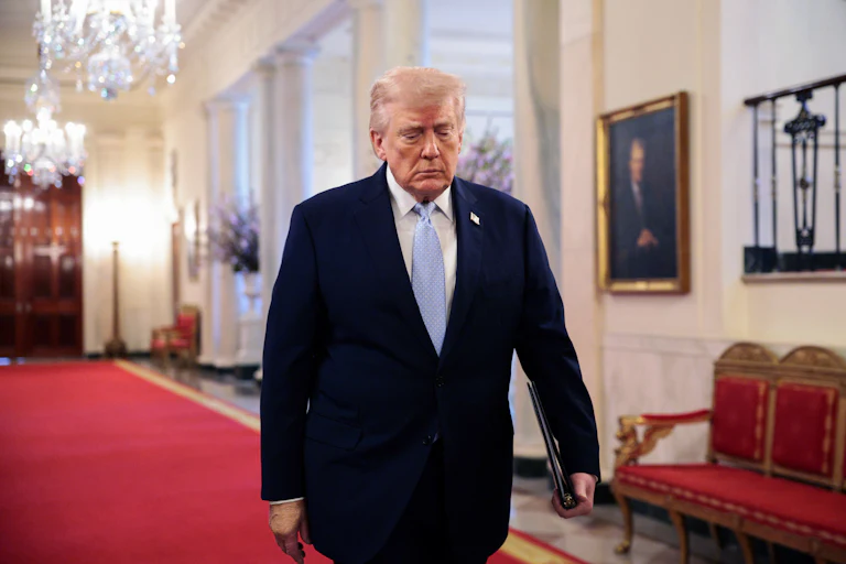 Trump walks on red carpet through the East Room of the White House wearing a suit and light blue tie, with chandeliers visible above and behind him.