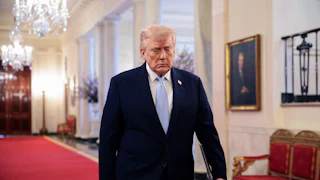 Trump walks on red carpet through the East Room of the White House wearing a suit and light blue tie, with chandeliers visible above and behind him.