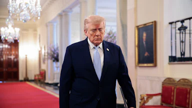 Trump walks on red carpet through the East Room of the White House wearing a suit and light blue tie, with chandeliers visible above and behind him.