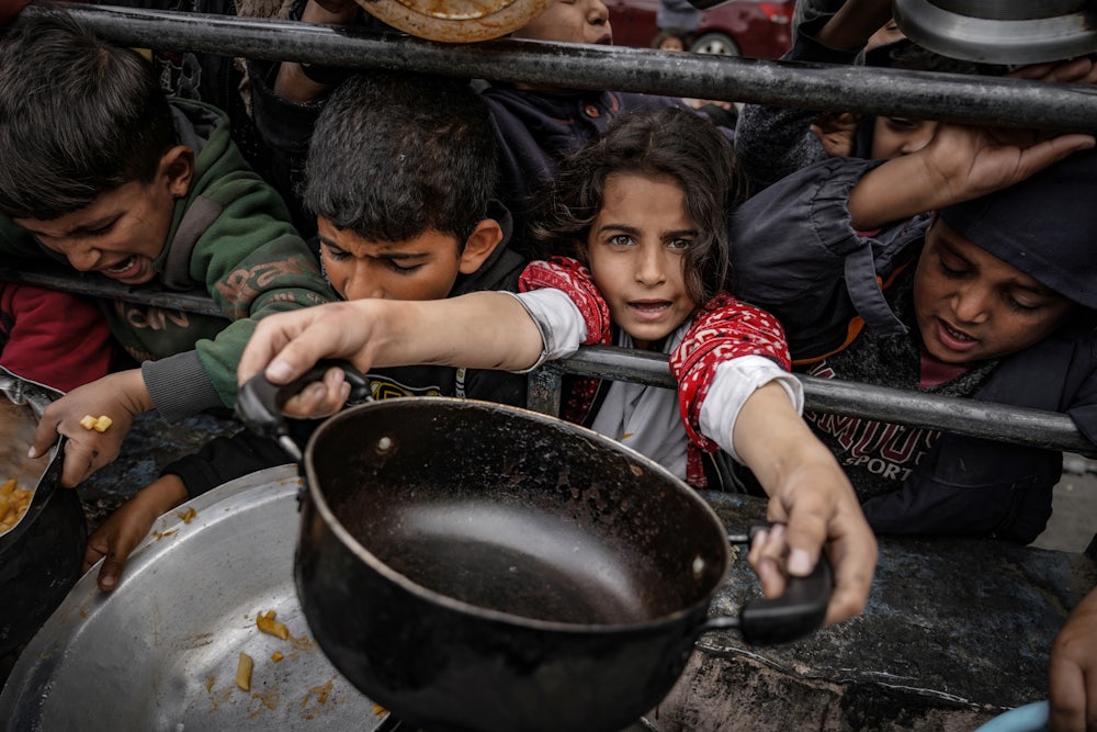 Palestinian children, holding empty pots, wait in line to receive food prepared by volunteers for Palestinian families displaced to Southern Gaza due to Israeli attacks, between rubbles of destroyed buildings in Rafah, Gaza on February 10, 2024.
