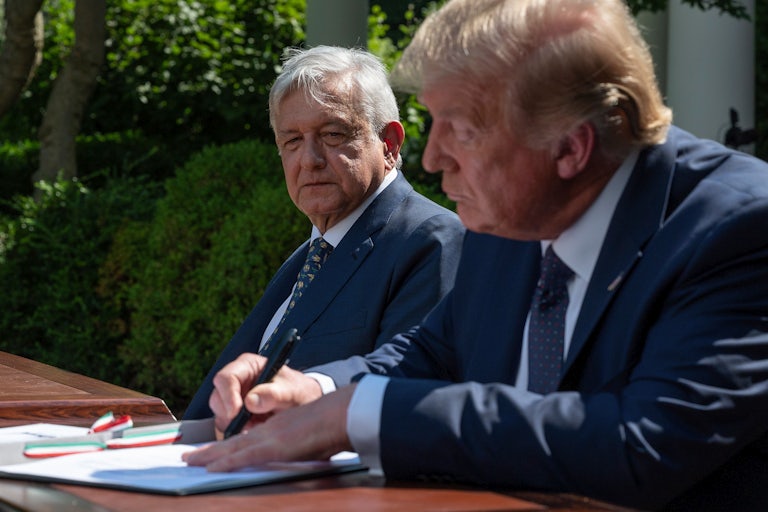 President López Obrador looks at President Trump, both seated at a desk as Trump signs a document.