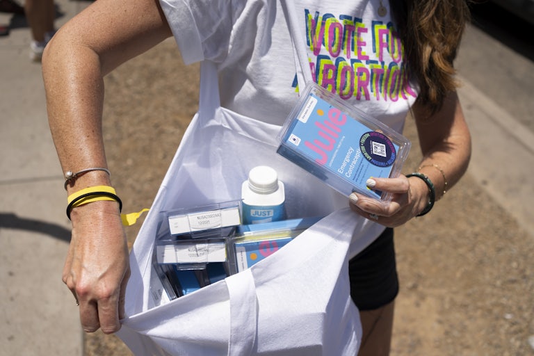 A person shows the contents of a bag full of emergency contraception while wearing a shirt that reads "Vote for Abortion"