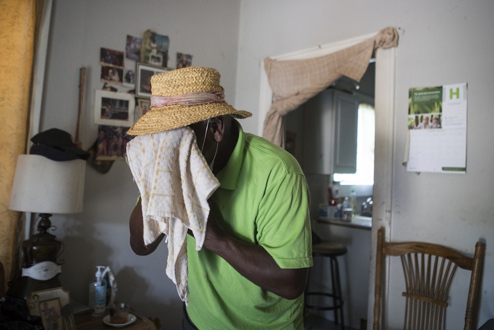 A man wearing a straw hat stands indoors and holds a towel to his face.