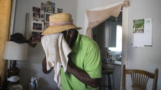 A man wearing a straw hat stands indoors and holds a towel to his face.