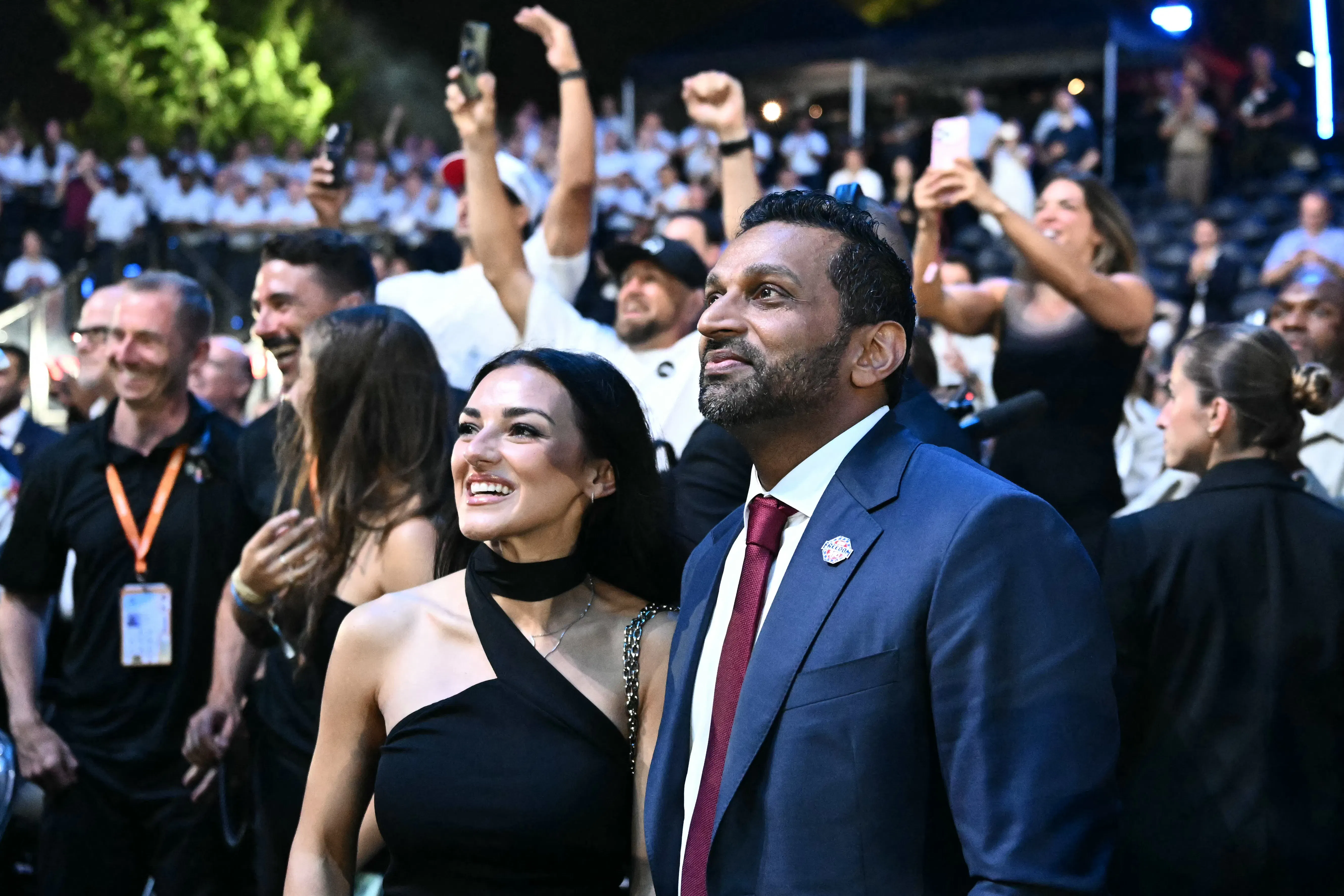 Alexis Wilkins stands on the right in a black top next to Kash Patel in a blue suit at night, outdoors, with a crowd of people behind them, some holding up phones to capture video.