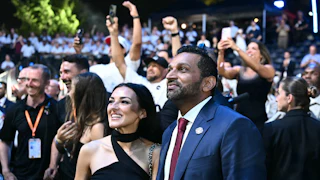 Alexis Wilkins stands on the right in a black top next to Kash Patel in a blue suit at night, outdoors, with a crowd of people behind them, some holding up phones to capture video.