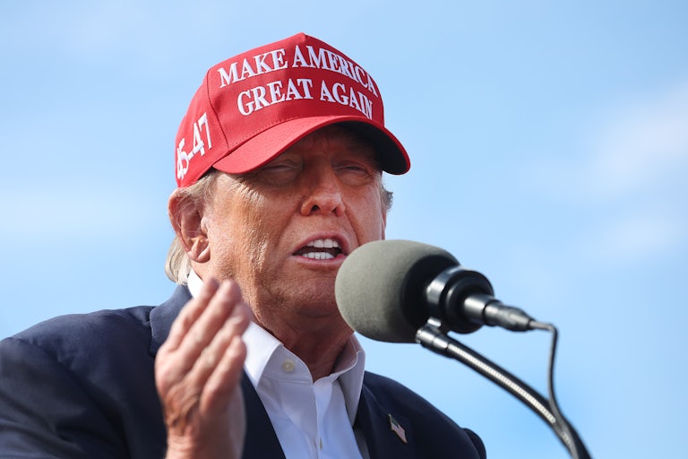 Donald Trump wears a red Make America Great Again cap and yells into a mic while squinting